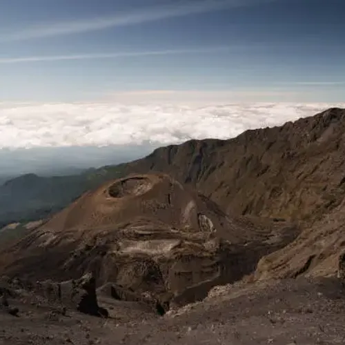 View of Mount Meru showcasing its volcanic crater, rocky ridges, and dramatic high-altitude landscape above a blanket of clouds.